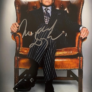 Może przedstawiać: Podpisane zdjęcie mężczyzny w garniturze w paski, siedzącego w brązowym skórzanym fotelu. Ma na sobie wzorzysty krawat i czarne buty. Podpis jest srebrnym atramentem.