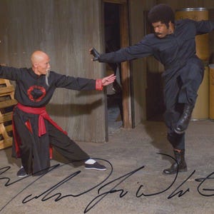 May include: A vintage photograph shows two men in martial arts stances. One is in a black and red martial arts uniform, the other in a black jumpsuit. The background features wooden pallets and barrels, with signatures at the bottom.