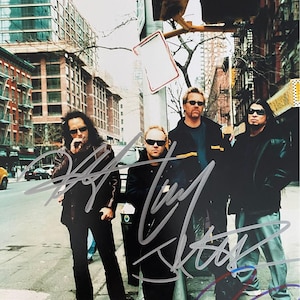 Puede incluir: Fotografía en color de una banda de rock posando en una calle de la ciudad. Los miembros de la banda llevan gafas de sol y ropa oscura. La imagen está firmada por los miembros de la banda.