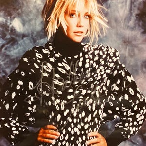 Puede incluir: Una mujer con cabello rubio lleva un cuello alto negro y una blusa con estampado en blanco y negro. La blusa tiene un diseño de lunares. La mujer posa con las manos en las caderas. El fondo es un degradado azul y gris.