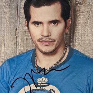 May include: A portrait of a man with dark hair and a mustache, wearing a blue t-shirt with a graphic design and a silver chain necklace. The background is a wooden wall.