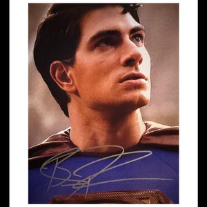 May include: A framed portrait of a man with dark hair, blue eyes, and a determined expression. He is wearing a blue and red costume with a brown cape. The image is signed in silver ink.