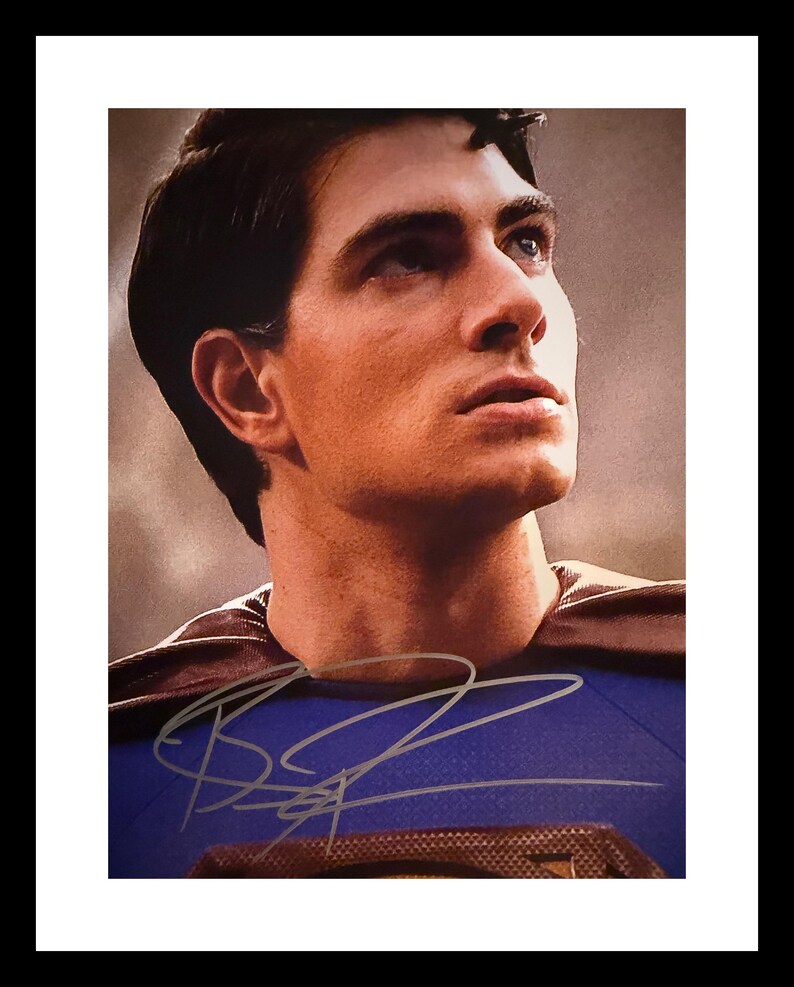 May include: A framed portrait of a man with dark hair, blue eyes, and a determined expression. He is wearing a blue and red costume with a brown cape. The image is signed in silver ink.
