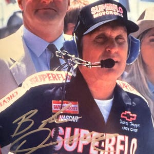 May include: A man wearing a black Superflo Motor Oils hat and uniform with the text "SUPERFLO MOTOR OILS" and "Winston NASCAR". The uniform has a signature and the name "Harry Hegge, Crew Chief". The image is a close-up of the man.