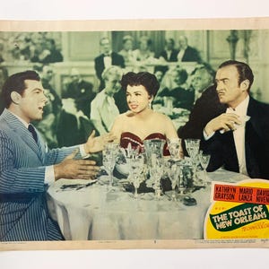Pode incluir: Cartaz de filme vintage de "The Toast of New Orleans" com atores sentados a uma mesa com copos. O cartaz tem uma faixa amarela e verde com o título do filme e os nomes do elenco. A cena passa-se num restaurante.