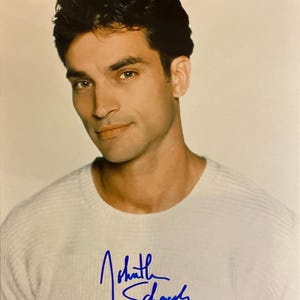May include: A portrait of a man with dark hair and a light-coloured sweater. The man is looking towards the camera. The sweater has a ribbed texture. The image includes a blue signature on the sweater.