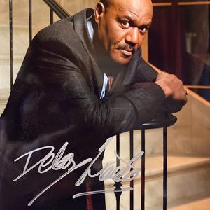 Puede incluir: Un retrato de un hombre con un traje oscuro, apoyado en una barandilla de escalera. La imagen presenta un autógrafo en escritura blanca. El hombre tiene la cabeza calva y un bigote, y mira directamente al espectador.