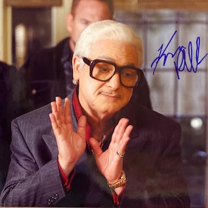 Op de afbeelding: Een gesigneerde foto van een man met wit haar, met een grote, donkeromrandde bril, een rood overhemd met kraag en een grijs pak met patroon. De man gebaart met zijn handen en een handtekening is zichtbaar in blauwe inkt.