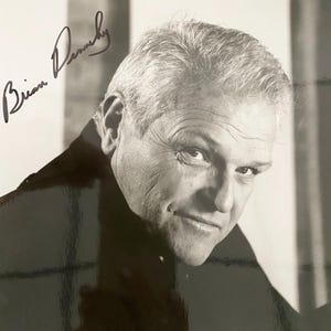 May include: Black and white photograph of a man with gray hair, looking towards the viewer. The image includes the handwritten signature "Brian Dennehy" in black ink.
