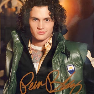 May include: A portrait of a person with curly dark hair wearing a green puffer vest over a plaid shirt and a floral print shirt. The vest has a small patch with a mountain and sun design. The image includes a signature in gold ink.