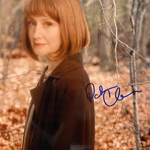 May include: A portrait of a woman with auburn hair and a brown coat, set against a blurred outdoor backdrop. The image includes a blue signature in the upper right corner.