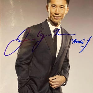 May include: A portrait of a man in a dark gray suit, white shirt, and black tie. The image includes a handwritten signature in blue ink. The man is smiling and looking at the camera.