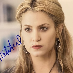 May include: Close-up of a woman with long blonde hair, wearing earrings and a necklace. Her lips are a deep red, and her eyes are brown. A blue signature is visible in the corner.