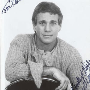 May include: A black and white photograph of a person with short hair wearing a textured knit cardigan over a button-up shirt. The image features handwritten ink inscriptions that read For Raul in the top left and Kindest Wishes Ryan O'Neal in the bottom right.