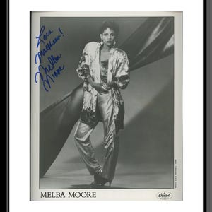 Puede incluir: Foto en blanco y negro de una mujer con una chaqueta larga y fluida con estampado y pantalones. Está de pie frente a un telón de fondo de tela drapeada. La foto está firmada "Love Matthew Melba Moore".