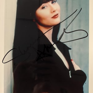 Könnte beinhalten: Signiertes Foto einer Frau mit langen schwarzen Haaren und einem schwarzen Blazer. Die Frau lächelt, mit rotem Lippenstift und Nagellack. Das Autogramm ist in schwarzer Tinte.