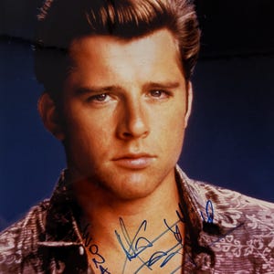 May include: A portrait of a man with light brown hair styled back. He is wearing a patterned button-down shirt. The image has a blue background and a signature in blue ink.
