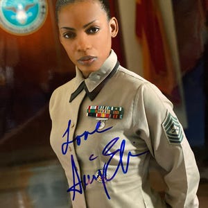Puede incluir: Retrato de una persona con uniforme militar beige, adornado con cintas, una insignia y una insignia de sargento. El uniforme está firmado con tinta azul. Un emblema circular es visible en el fondo.