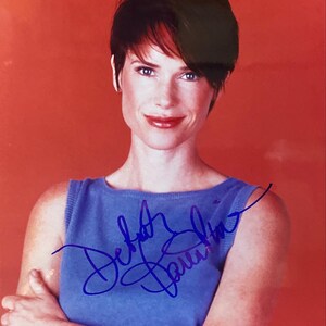 Puede incluir: Fotografía firmada de una mujer con pelo corto y oscuro y una parte superior azul. La firma, en tinta azul, está en la parte delantera de la parte superior. El fondo es de color rojo anaranjado sólido.