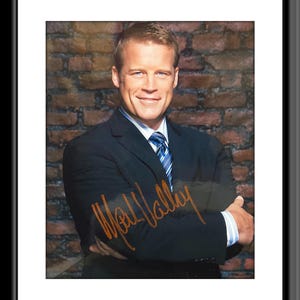 May include: A man in a dark blue suit with a light blue patterned tie, standing in front of a brick wall. He is smiling and looking at the camera. His signature is written in orange ink below his image.
