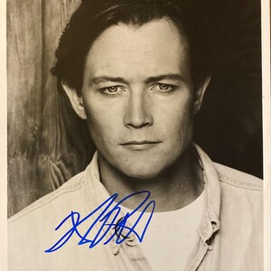 Op de afbeelding: Zwart-wit portret van een man met donker haar, gekleed in een lichtgekleurd overhemd. De afbeelding heeft een handtekening in blauwe inkt over het onderste gedeelte.