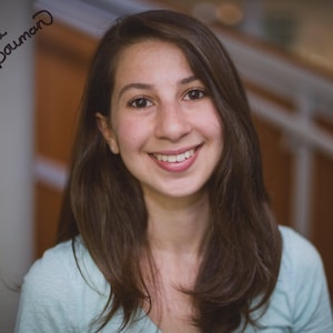 May include: A smiling person with long brown hair and a light blue shirt. The image includes the handwritten text "Katie Bauman" in the upper left corner. The background is blurred, with hints of stairs and a neutral wall.