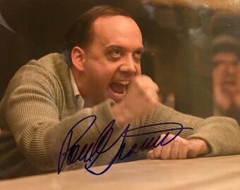 May include: A signed photograph featuring a man with an open mouth and clenched fist. The man is wearing a light gray sweater and is leaning on a table. The signature is in blue ink.
