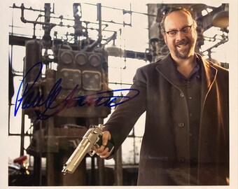 May include: A signed photograph featuring a man in a dark coat holding a silver handgun. The background includes industrial machinery. The signature is in blue ink. The man is wearing glasses and smiling.