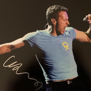 May include: A light blue short-sleeve t-shirt with a yellow logo on the chest pocket, worn by a person singing into a black microphone. The image features a white handwritten autograph in the lower left corner against a dark background.