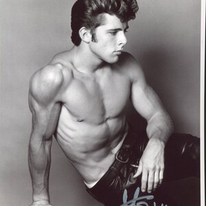 May include: A black and white photograph of a person with a muscular build, styled with a pompadour hairstyle, wearing dark leather trousers with a belt. The image features a handwritten signature in the bottom right corner in a light grey ink.