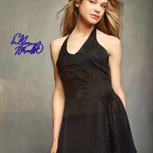 May include: A woman in a black halter dress with a polka dot pattern. The dress has a fitted bodice and a flared skirt. The woman has long blonde hair and is wearing earrings. The image includes a signature in blue ink.