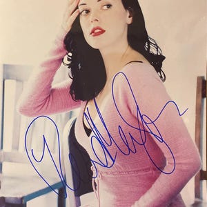 May include: A photograph of a person wearing a light pink sweater and dress, with dark hair and red lipstick. The image has a blue signature across the front. The person is seated, with one hand on their head.