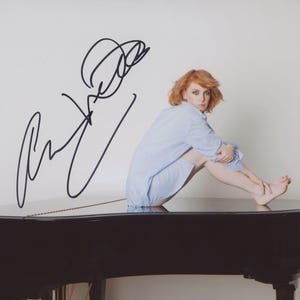 Puede incluir: Fotografía de una persona pelirroja sentada en un piano negro. La persona lleva una prenda azul claro y está descalza. El piano tiene un acabado brillante. Una firma negra está en la esquina superior izquierda de la imagen.