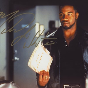 May include: A signed photograph of a man in a black leather jacket holding a document marked "TOP SECRET." The image has a gold signature in the upper left corner. The man is looking off to the side.