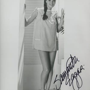 Puede incluir: Fotografía en blanco y negro de una mujer con un vestido corto de color claro con un bolsillo. Sonríe y hace el signo de la paz. La imagen está firmada "Samantha Eggar."