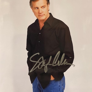 Puede incluir: Fotografía firmada de un hombre con una camisa negra y vaqueros azules. La firma es con tinta plateada. El fondo es blanco liso.