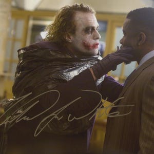 May include: A signed photograph depicting two men in a film scene. One man, with white face paint and red lipstick, is touching the other man's face. The photograph is signed in silver ink.