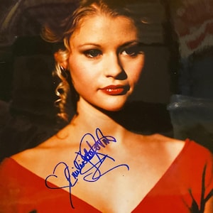 May include: A portrait of a woman with blonde hair and red lipstick, wearing a red dress. The image is signed in blue ink. The woman is looking towards the camera.
