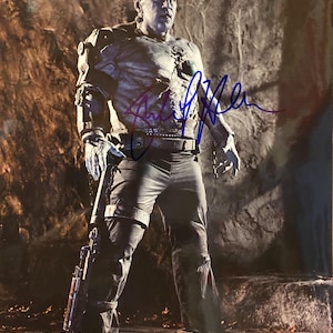 May include: A full-length image of a character in a monster costume, with gray skin and a scarred face. The character wears a dark outfit with metal accents and holds a weapon. The image is signed in blue ink.
