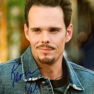 May include: A portrait of a man with fair skin, blue eyes, and a mustache. He wears a denim jacket over a dark shirt. The jacket has visible stitching. The image includes a blue signature on the jacket.