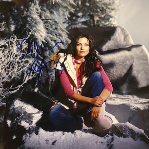 May include: A woman in a winter scene, wearing a pink sweater, a white vest with a fur-lined hood, and blue jeans. She is sitting on a snow-covered rock, with a snowy background and a signature in blue ink.