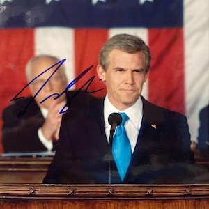 May include: A man in a dark suit and a light blue tie stands at a podium with a microphone. The background features an American flag. An autograph is visible in the upper left corner.