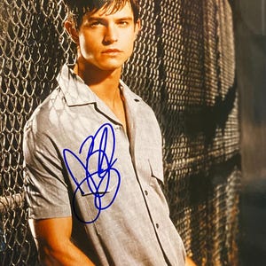 May include: A light gray short-sleeved button-down shirt with a pocket on the chest. The shirt is signed in blue ink. The background features a chain-link fence.