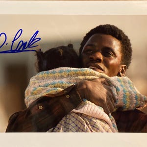 Puede incluir: Fotografía firmada que muestra a un hombre abrazando a un niño. El hombre, de piel oscura y pelo corto, lleva una camisa a cuadros. El niño está envuelto en una manta colorida y estampada. La firma "D. Locke" es visible en tinta azul.