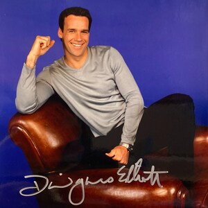 Puede incluir: Fotografía firmada de una persona sonriente con una camisa gris de manga larga y pantalones negros. La persona está sentada en un sillón de cuero marrón sobre un fondo azul. Firma "Dwigans Elliott".