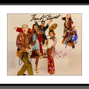 May include: A framed photograph of a band with five members, all wearing 1960s-style clothing. The band members are holding guitars and a microphone. The photograph is signed by all five band members.