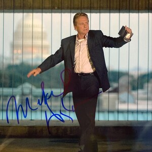 Może przedstawiać: Autografowane zdjęcie mężczyzny w ciemnym garniturze, białej koszuli i czarnym pasku. Mężczyzna trzyma mały przedmiot i ma poważny wyraz twarzy. Tło obejmuje rozmyty budynek i podpis niebieskim tuszem.