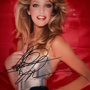 Puede incluir: Retrato de una mujer con cabello rubio y ojos azules, vestida con un top plateado, un cinturón plateado y vaqueros azul oscuro. El fondo es de color rojo liso. La imagen tiene un autógrafo en tinta negra.