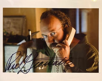 May include: A signed photograph featuring a man with glasses holding a telephone receiver to his ear. The man is wearing a green shirt and a watch. The signature is in black ink.