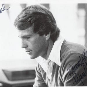 May include: A black and white photograph of a person with short hair wearing a collared shirt under a V-neck sweater. The image features handwritten ink signatures in the top left and bottom right corners, including the name Ryan O'Neal.